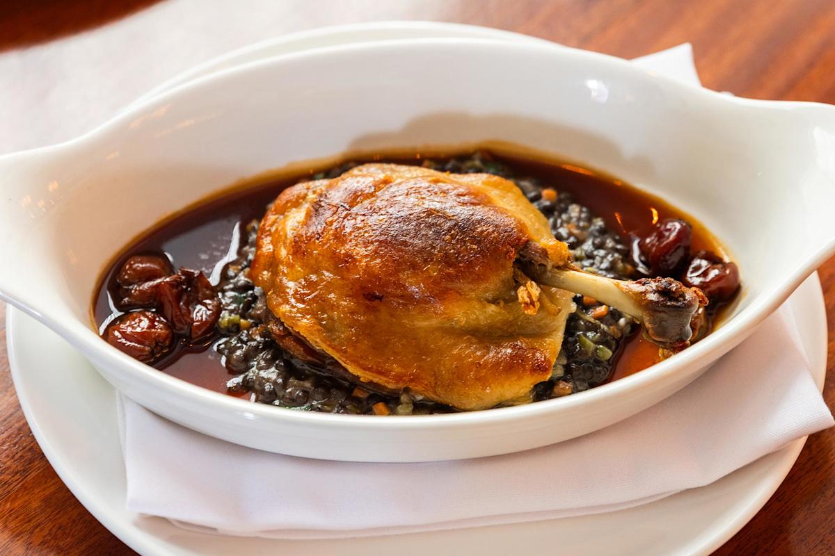 A golden-brown roasted duck leg served on a bed of lentils with a dark, rich sauce and garnished with cherries in a white oval dish atop a folded white napkin.