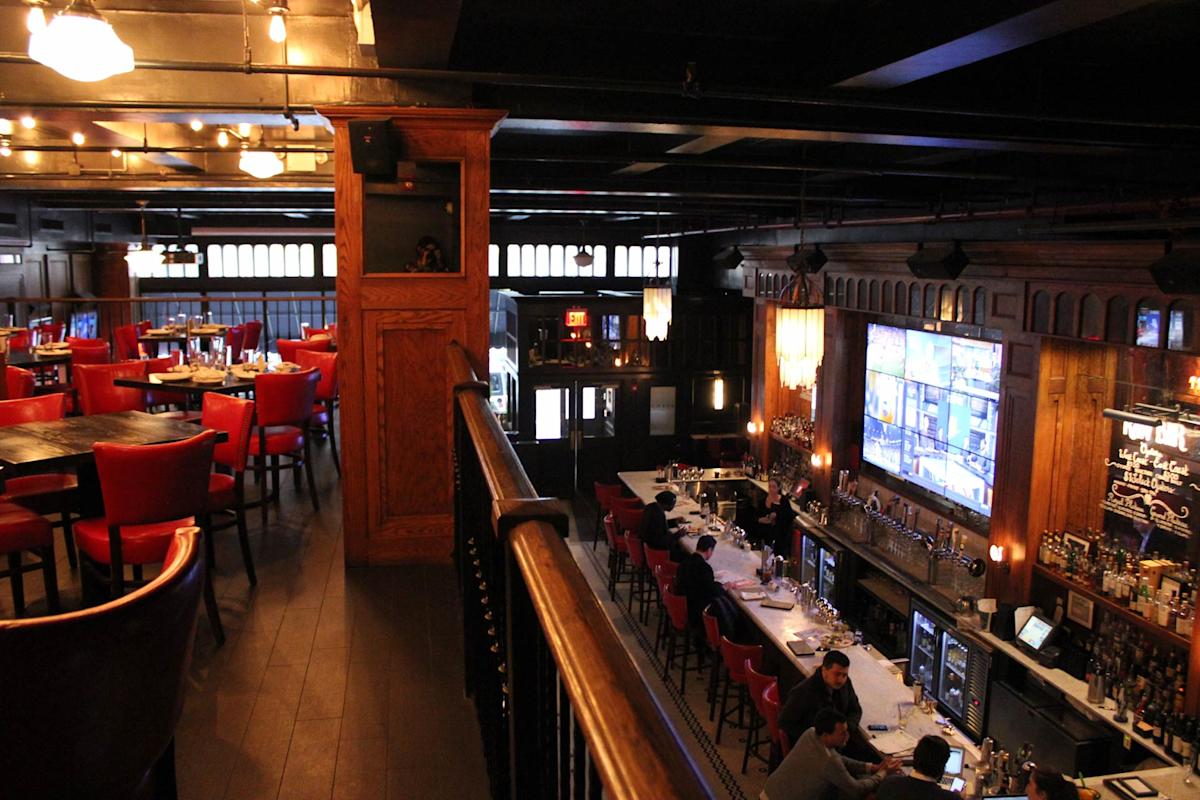 A view of a stylish bar and restaurant with high ceilings, red chairs, wooden accents, and large TVs above the bar. People are seated at the bar and tables, creating a lively, inviting atmosphere.