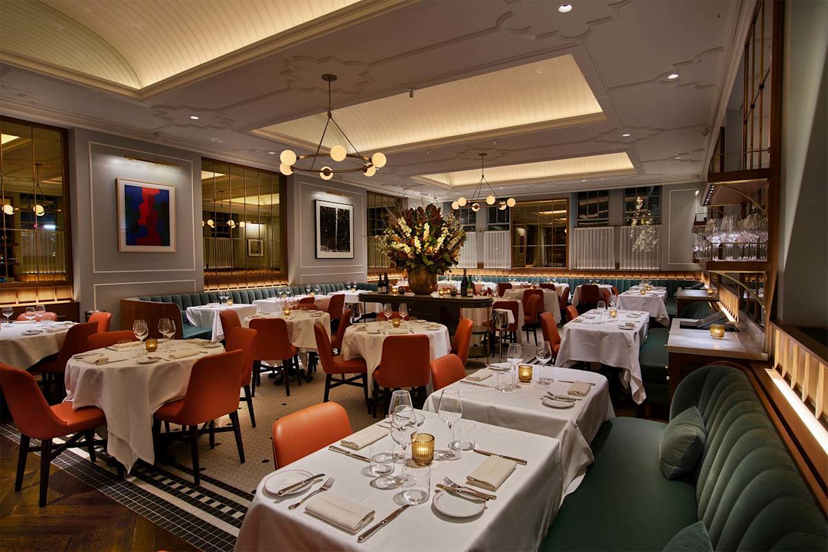 Elegant restaurant interior with neatly set tables, orange chairs, green banquettes, soft lighting, modern art on the walls, and a large floral centerpiece creating a warm, inviting atmosphere.