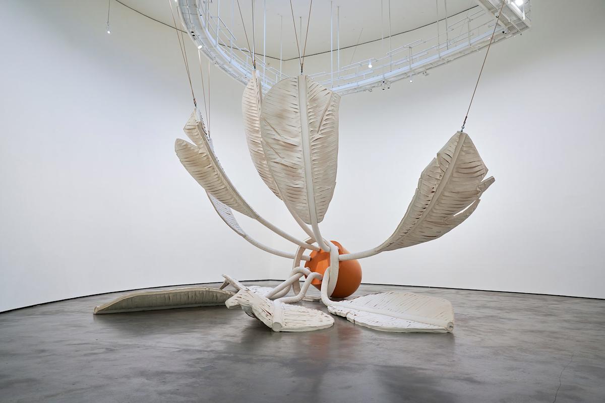 A large white sculptural installation resembling oversized leaves and stems radiates from an orange sphere, suspended and lying on a polished concrete floor in a minimal, curved white gallery space.