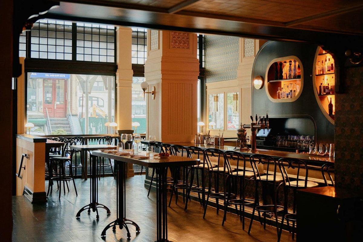 A cozy, stylish bar interior with wooden tables, black chairs, a long bar counter, and bottles displayed on rounded wall shelves. Large windows let in natural light, revealing a street view outside.