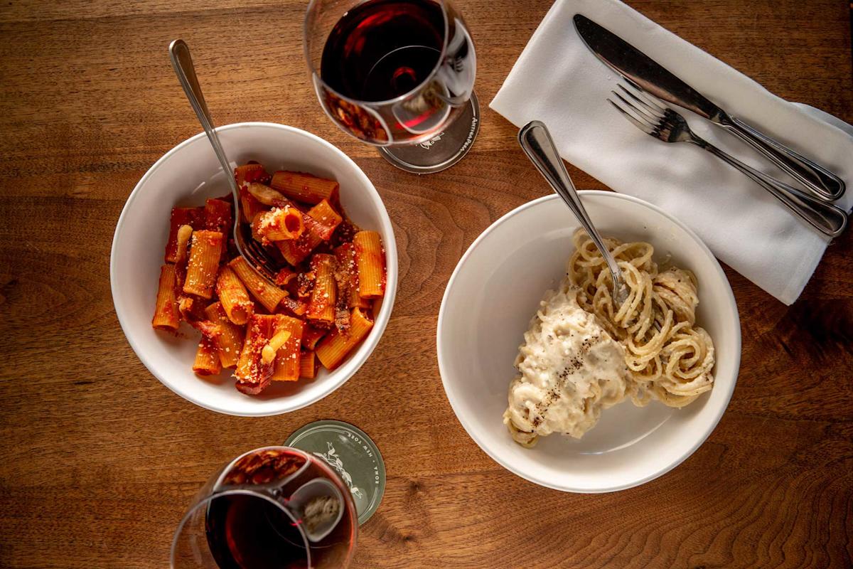 Two bowls of pasta, one with tomato sauce and the other with creamy white sauce, are on a wooden table with two glasses of red wine, a napkin, and silverware.