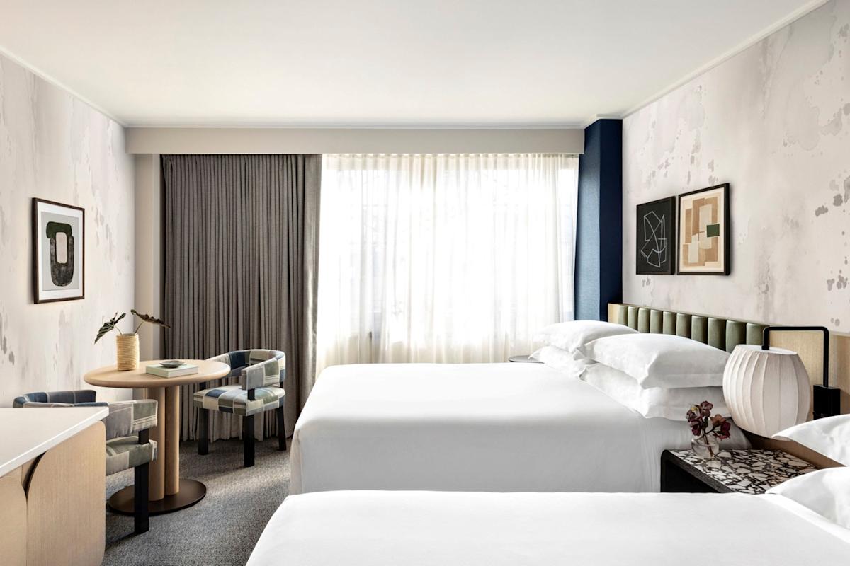 Bright hotel room with two double beds, a small round table with two chairs, a large window with sheer curtains, neutral-toned walls, framed abstract art, and a modern white lamp on the nightstand.