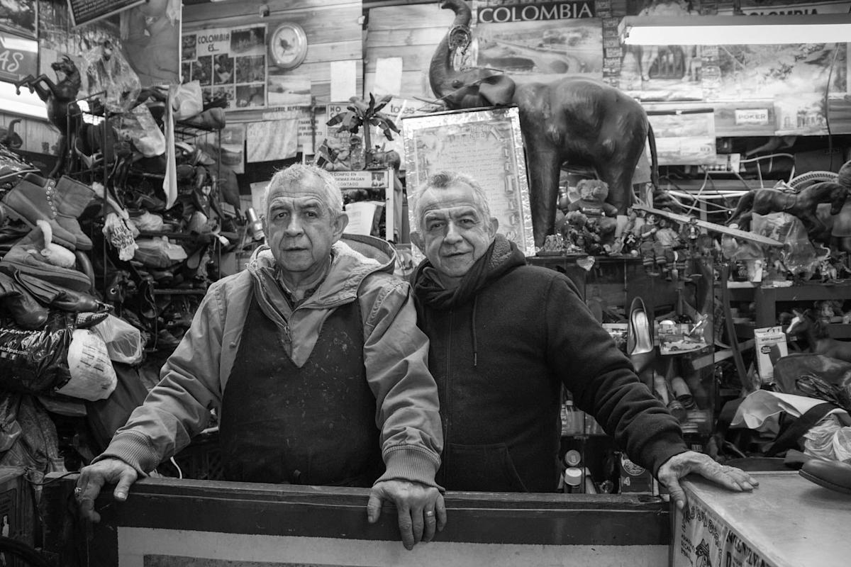 Two older men stand behind a cluttered counter in a shop filled with various items, including an elephant statue and posters. The black-and-white photo captures a cozy, nostalgic atmosphere.