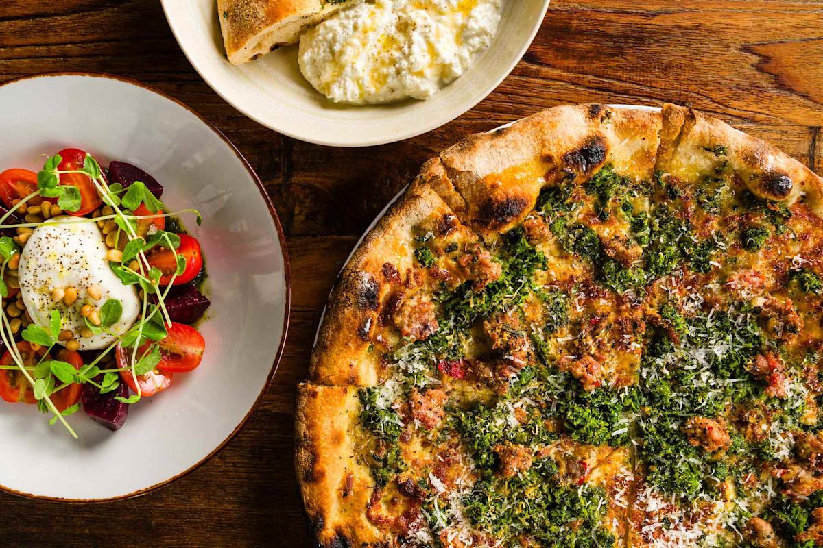 A wood-fired pizza topped with greens, herbs, and cheese sits beside a fresh salad with tomatoes, beets, burrata, and microgreens, and a bowl of creamy ricotta with bread on a rustic wooden table.