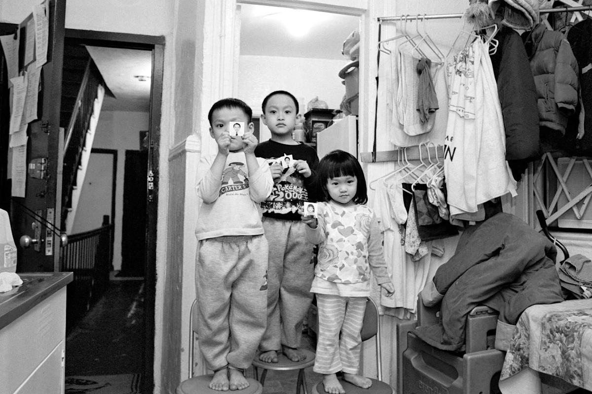 Three children stand in a small, cluttered room. Two hold toy figures while the third looks ahead. Clothes hang on a rack, and stairs are visible in the background. The atmosphere is casual and lived-in.
