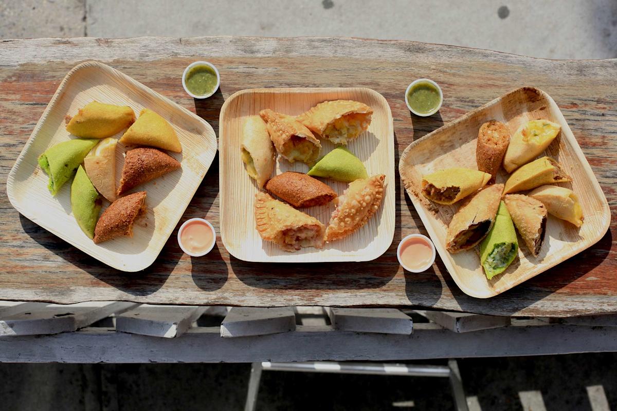 Three wooden trays on a rustic table, each filled with assorted empanadas and dipping sauces in small cups. The empanadas are golden brown, some with green or yellow hues, and the trays are arranged side by side.