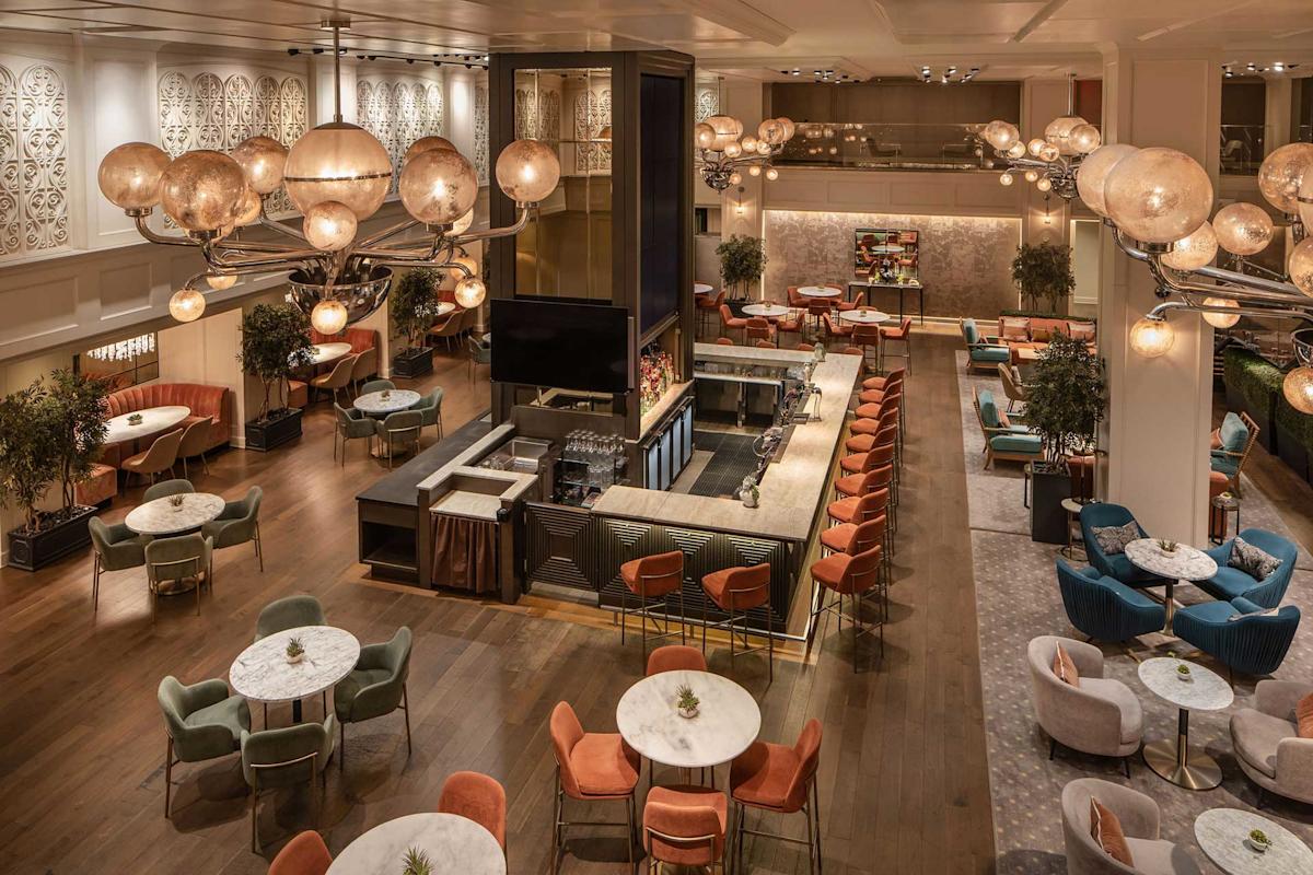 A spacious, elegant lounge with round tables, colorful velvet chairs and sofas, modern chandeliers with globe lights, a central bar area, and warm ambient lighting.