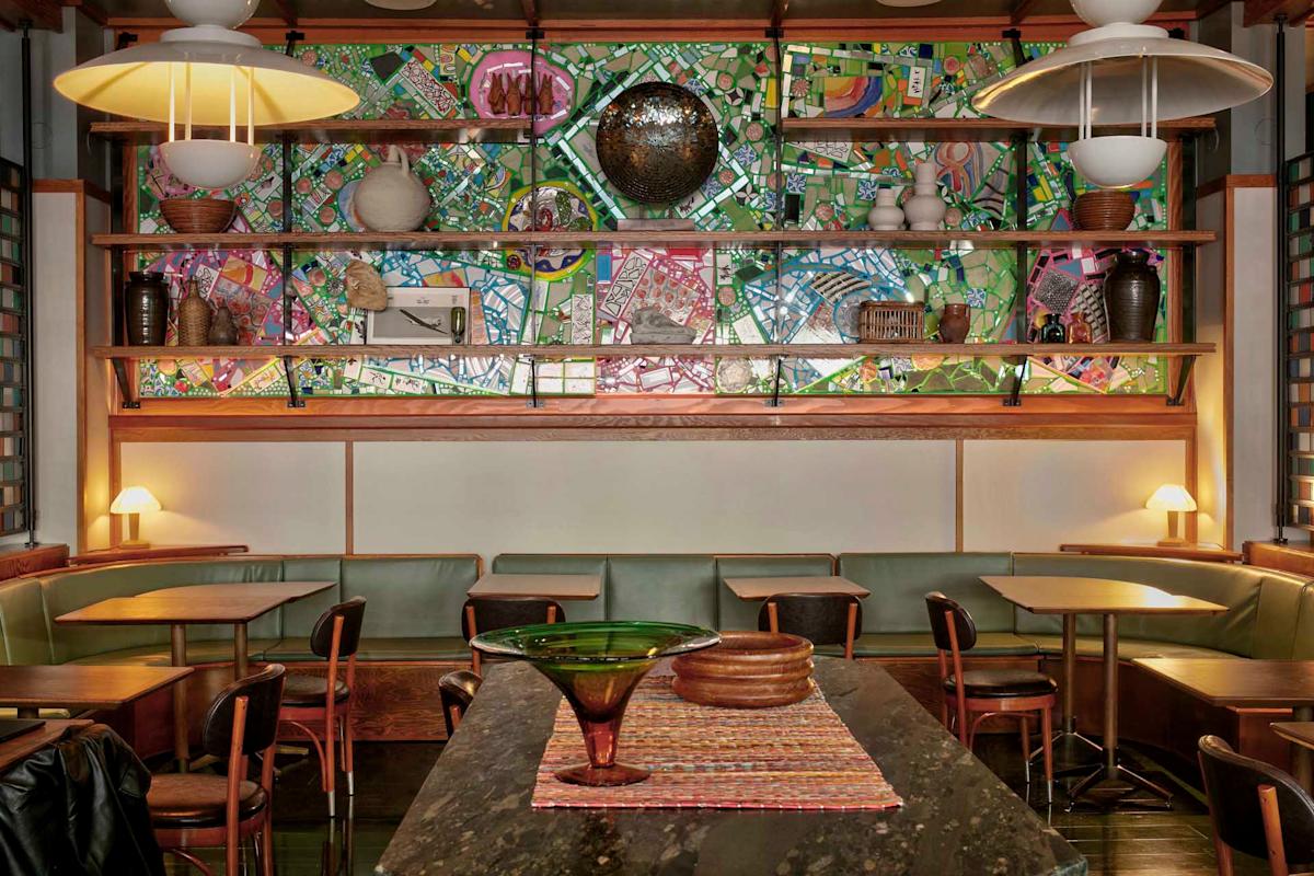 Cozy café with wooden tables and chairs, green cushioned benches, pendant lights, and shelves displaying vases and decor. A colorful, abstract mural decorates the back wall behind the shelves.