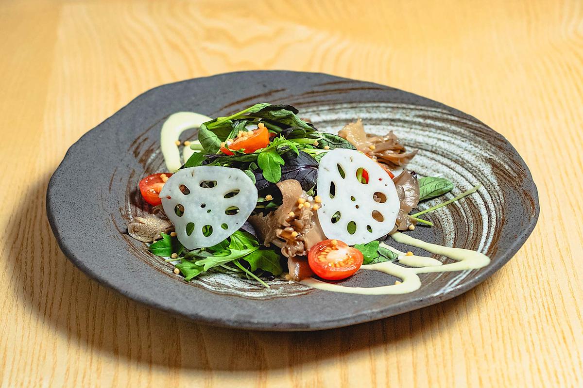 A black ceramic plate with a salad of mixed greens, cherry tomato halves, mushrooms, and thinly sliced lotus root, drizzled with a light dressing, on a light wooden table.