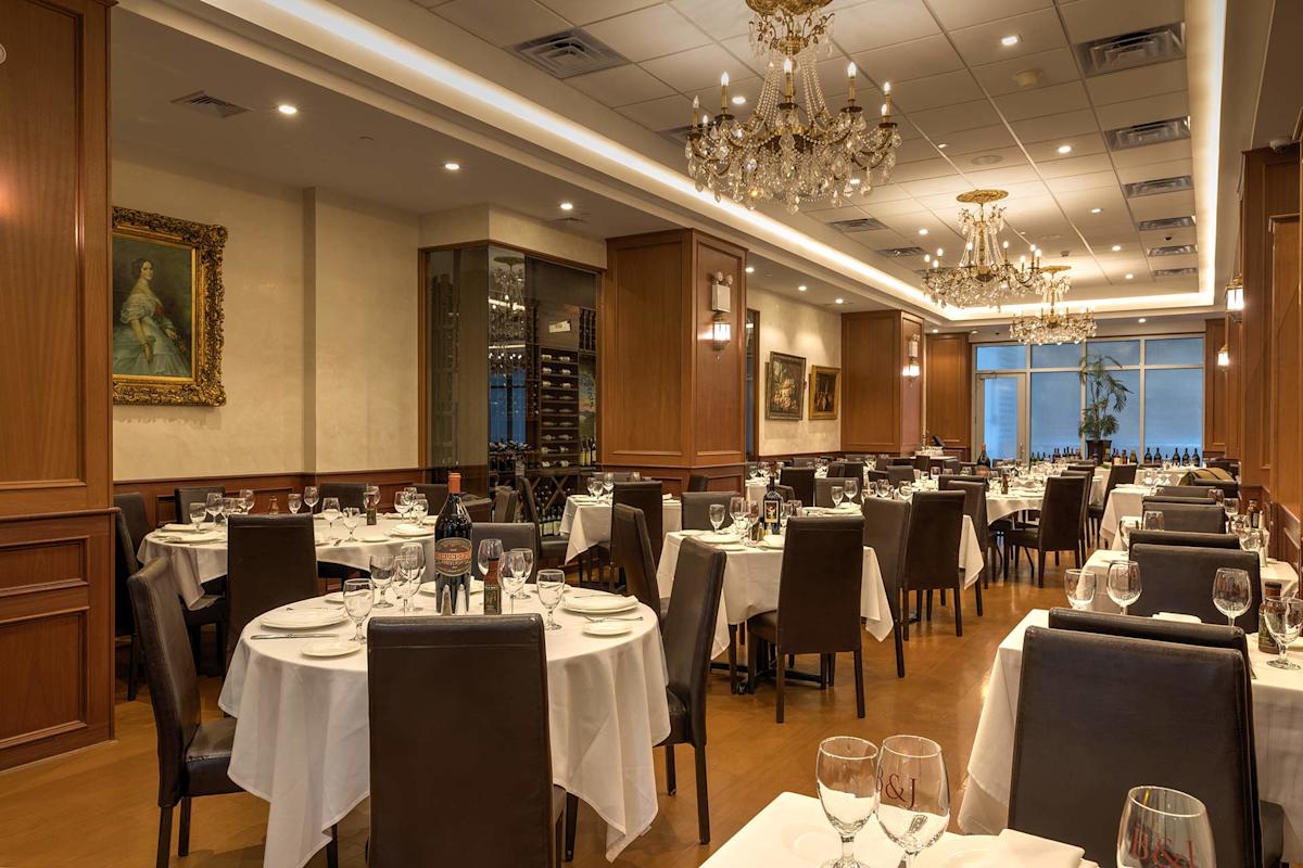 Elegant restaurant dining room with chandeliers, wooden paneling, and tables set with white tablecloths, glassware, and wine bottles. Framed artwork decorates the walls, creating a sophisticated atmosphere.