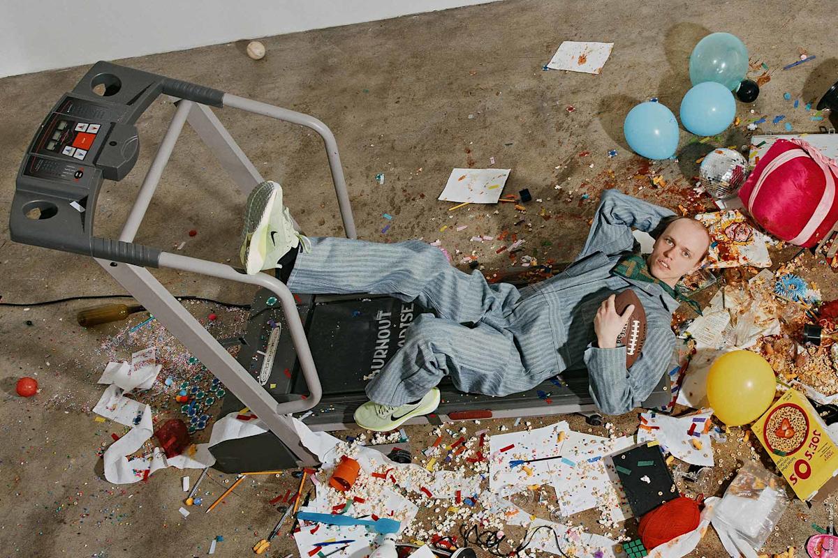 A man in striped pajamas lies on a treadmill, clutching a football, surrounded by a messy floor scattered with balloons, confetti, food, and party debris.