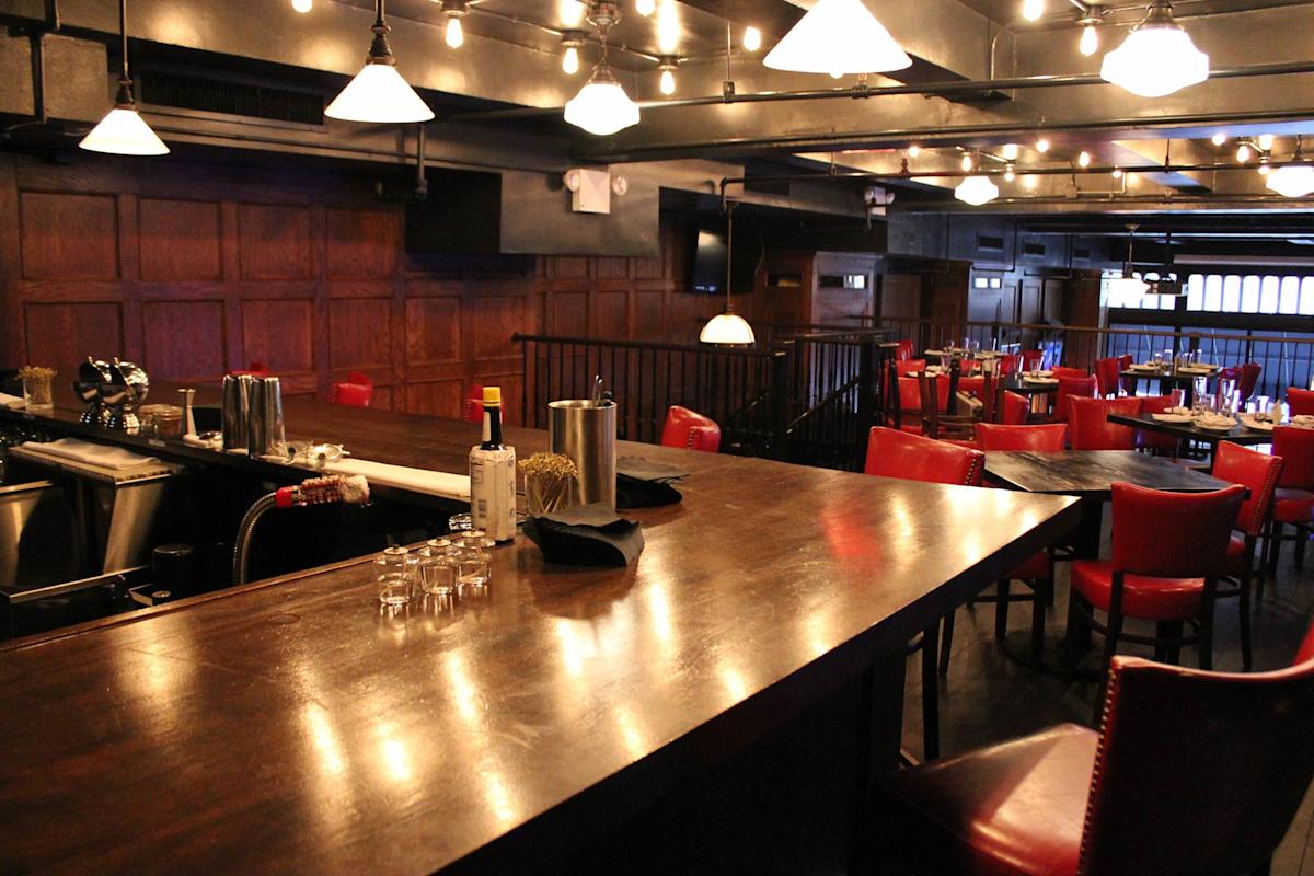 A dimly lit bar with wooden walls, a polished countertop, red chairs, and tables. Bottles and glasses are on the bar, and string lights hang from the ceiling, creating a cozy atmosphere.