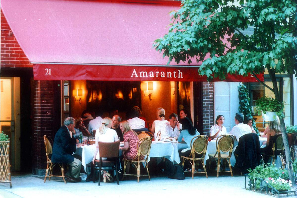 People dining outdoors at Amaranth restaurant under a red awning. Tables with white tablecloths are set on the sidewalk, with diners enjoying food and conversation on a sunny day.