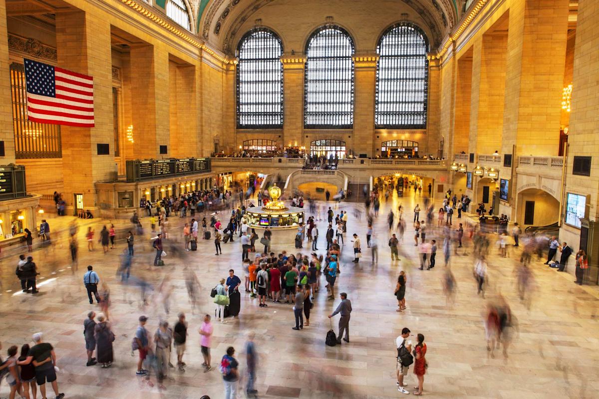 The main hall of Grand Central Terminal in New York City, filled with blurred crowds of people walking. Large arched windows and an American flag are visible. Natural light streams into the spacious interior.