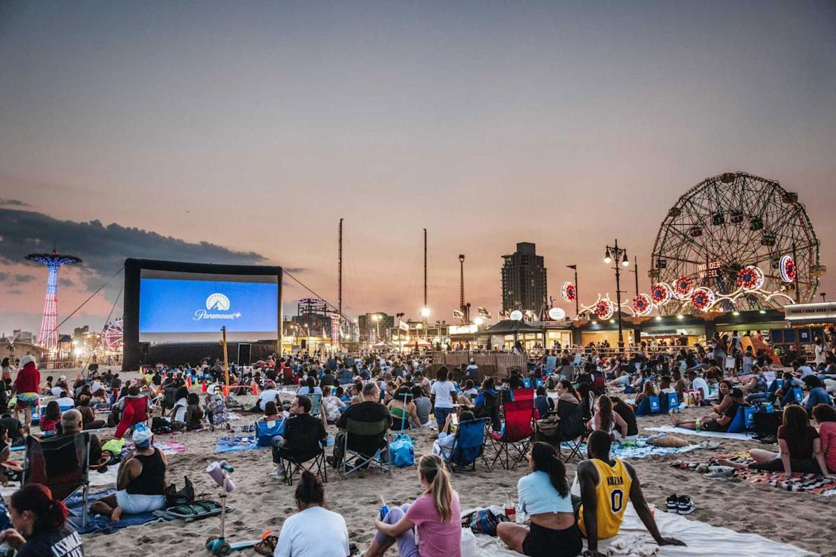 A large crowd sits on blankets and chairs on a beach at sunset, watching a movie on a big outdoor screen. Coney Island rides and bright lights are visible in the background.