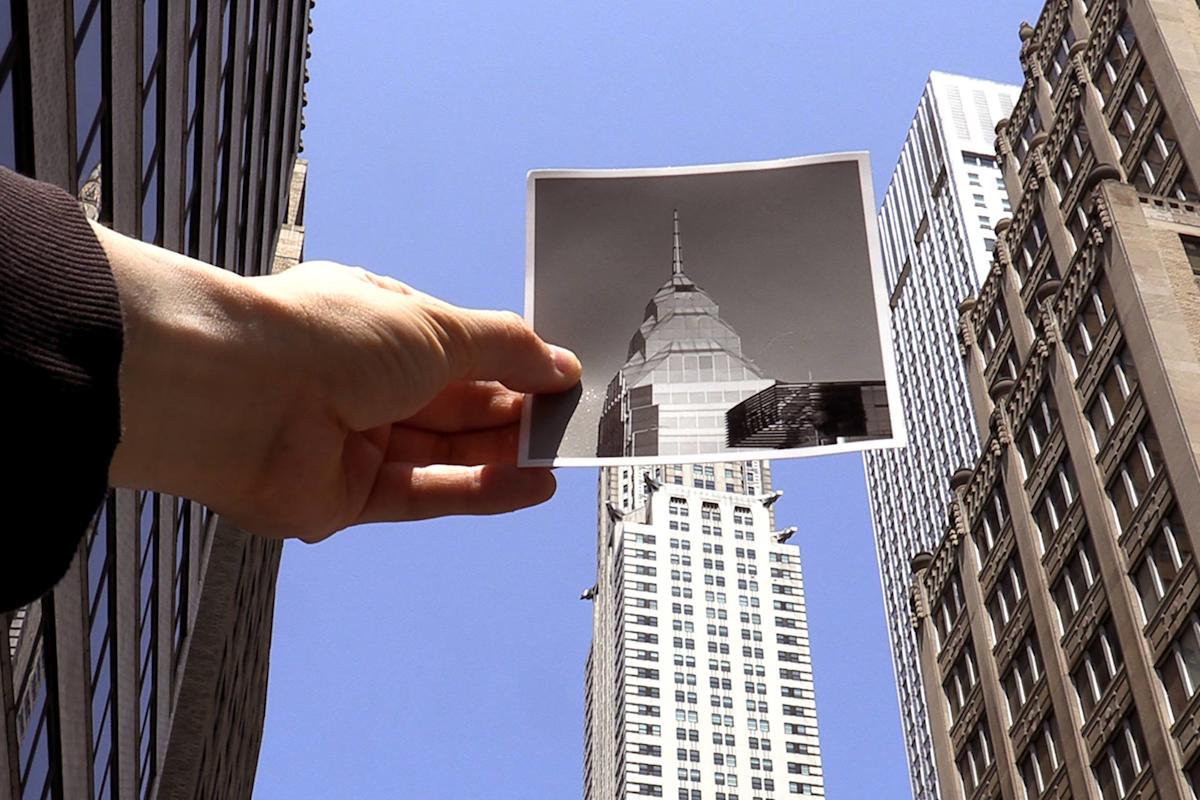 A hand holds an old black-and-white photo of a building, aligning it with the same modern skyscraper in the background, surrounded by other tall buildings against a clear blue sky.
