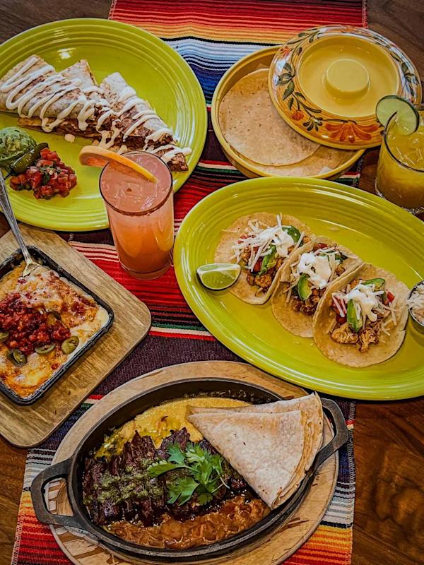A vibrant Mexican meal with tacos, tortillas, grilled steak with beans, cheesy dip, a dessert with cream, and two colorful drinks, all served on green plates atop a striped tablecloth.