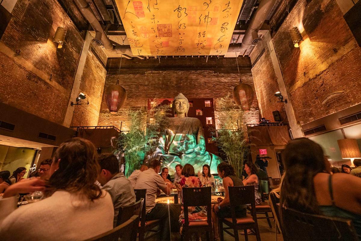 A warmly lit restaurant with high brick walls features a large Buddha statue as its centerpiece. Diners sit at tables around the statue, creating a lively, inviting atmosphere. Asian-inspired decor includes bamboo and hanging lanterns.
