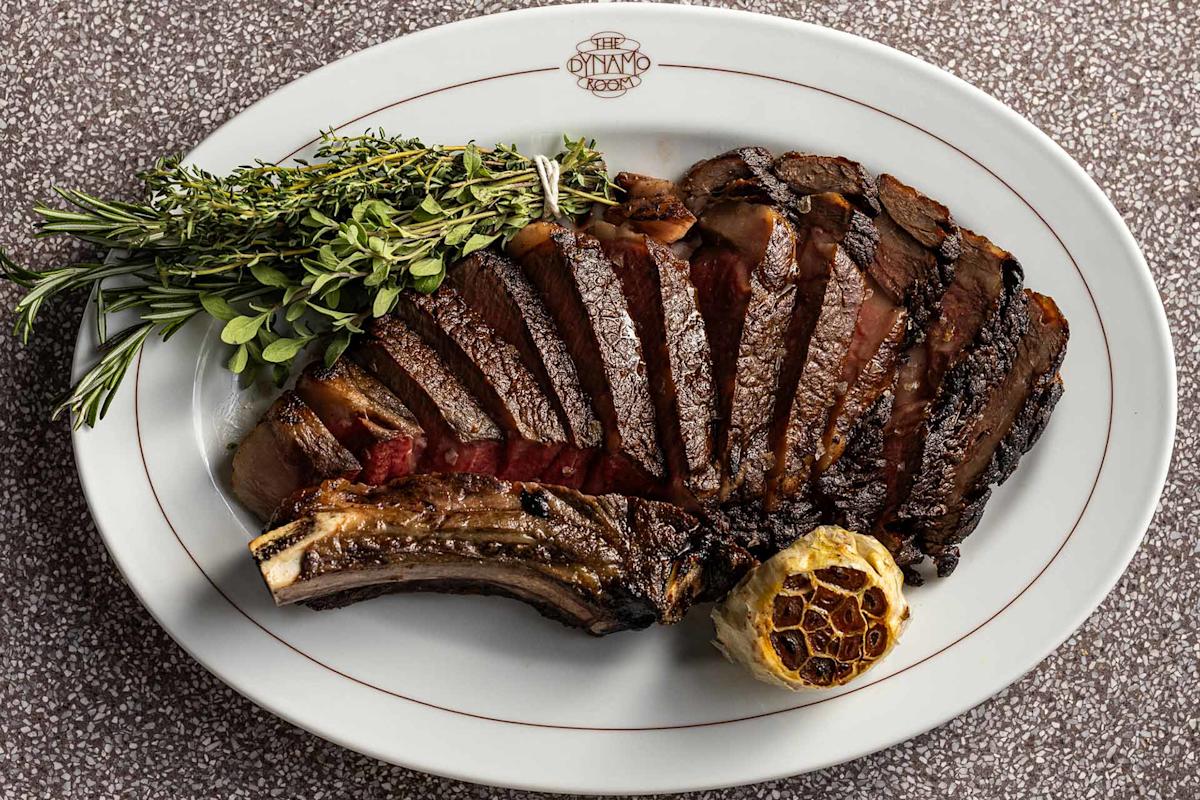 A sliced, grilled bone-in steak is arranged on a white oval plate with a roasted garlic bulb and fresh sprigs of herbs, including rosemary, on the side. The steak is cooked medium-rare and served on a patterned surface.