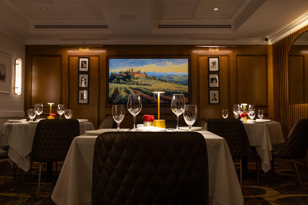 A dimly lit, elegant restaurant with tables set with white cloths, wine glasses, and a single red rose centerpiece. A large landscape painting and framed photos decorate the brown-paneled walls.