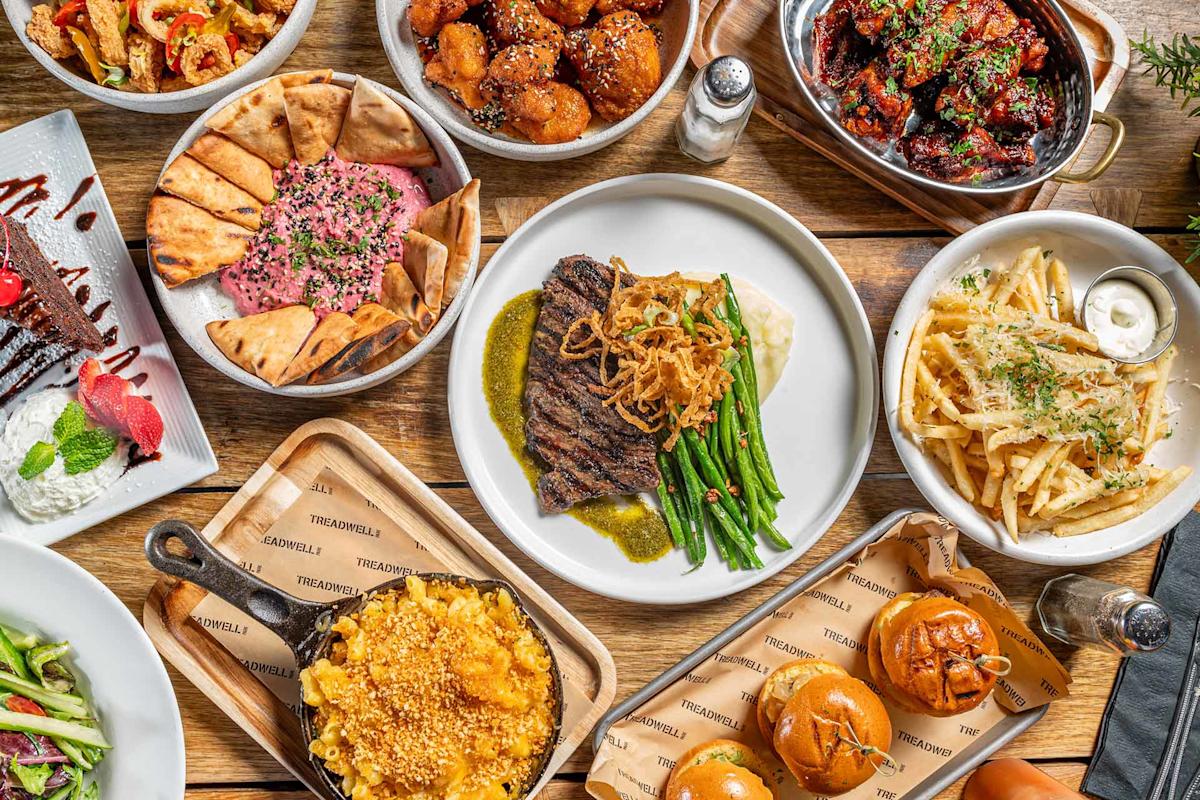 A variety of dishes on a wooden table, including steak with green beans, macaroni and cheese, fries with herbs, pita bread with dip, sliders, salad, wings, and chocolate cake dessert.