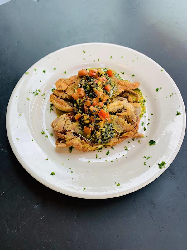 A white plate with sliced cooked meat topped with diced vegetables and herbs, garnished with chopped parsley, on a dark surface.