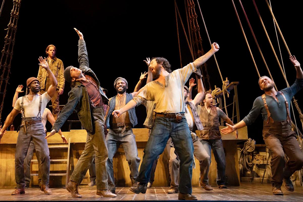 A group of performers energetically dance on a stage set resembling a ship's deck. They are dressed in period costumes and appear animated and joyful, with arms raised in various poses against a backdrop of ship rigging.