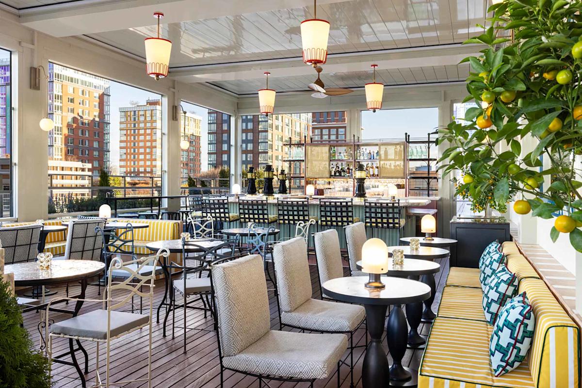 Chic rooftop restaurant with striped bench seating, cushioned chairs, marble tables, hanging lights, and city views through large windows. A lemon tree decorates the corner.
