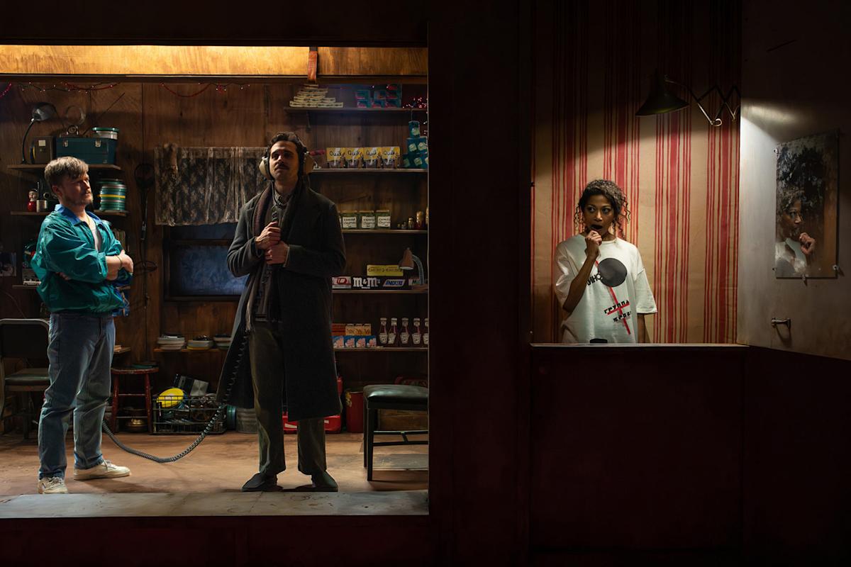 Two men stand in a cluttered shop, one in a teal jacket with arms crossed and the other in a long coat using a corded phone. In a separate, dimly lit room, a woman in a white shirt listens on another phone.