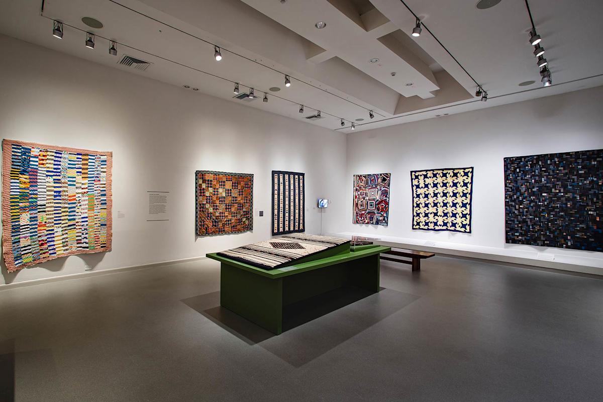 A modern art gallery room displaying colorful quilts on white walls and on a central green table, with spotlights illuminating the textiles and a bench for visitors.
