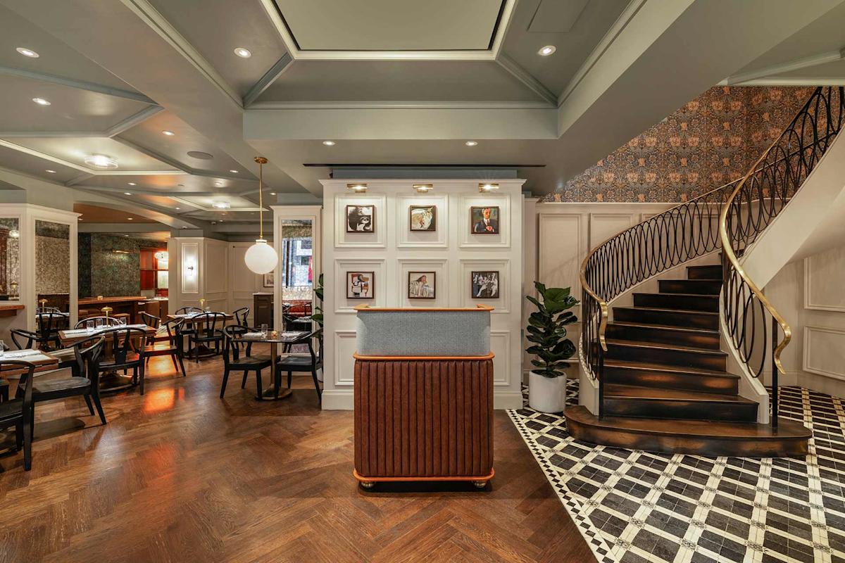 Elegant restaurant interior with a wooden host stand, framed photos on the wall, stylish wood staircase with a gold railing, patterned tile and wood flooring, and modern dining tables and chairs in the background.