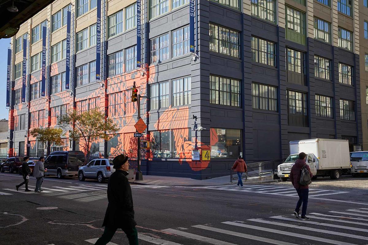 People cross a city street at a corner with a large industrial-style building featuring colorful geometric wall art. Parked cars and a white truck line the streets under sunny skies.