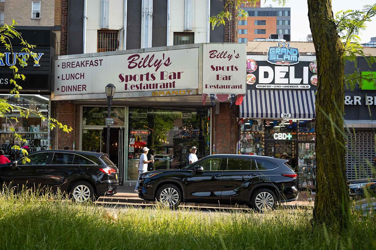 Street view of shops including Billy’s Sports Bar Restaurant and Grand Slam Deli, with cars parked in front, people walking on the sidewalk, and a grassy area and tree in the foreground.