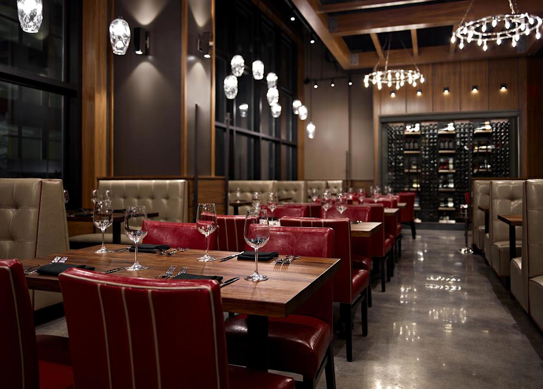 Modern restaurant interior with red leather chairs, wooden tables set with wine glasses and silverware, beige upholstered booths, wine rack, large windows, and decorative pendant lights. The space is empty and well-lit.