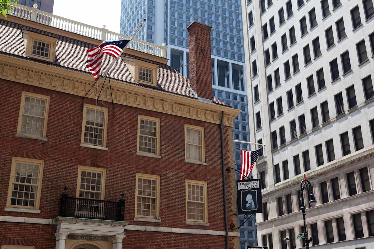 A historical brick building with American flags is surrounded by modern skyscrapers in a city. The building features colonial architecture with large windows and a prominent chimney.