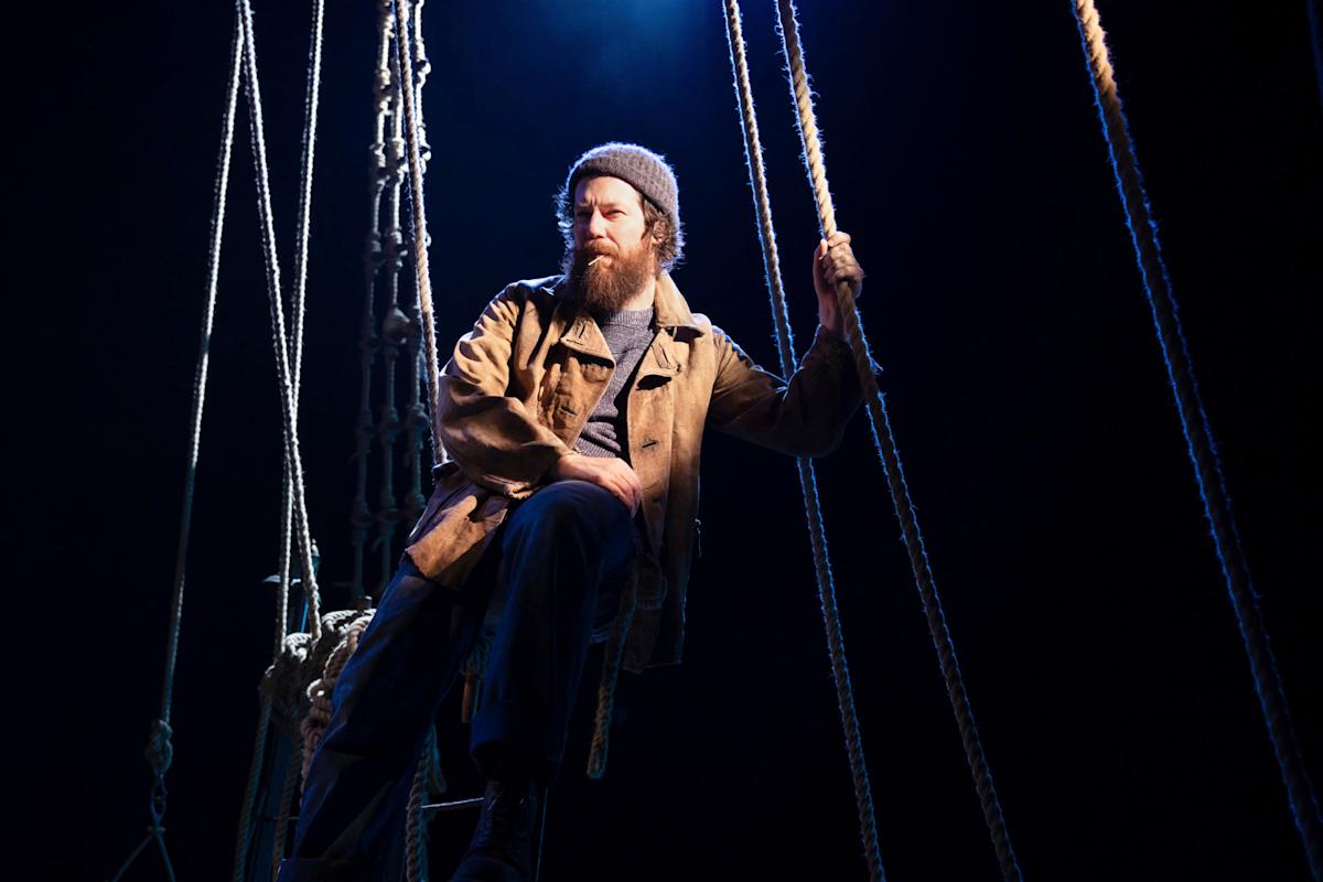 A person with a beard and beanie sits on a part of a ship, surrounded by ropes, under dramatic lighting on stage. They wear a brown jacket and blue pants, gazing into the distance. The setting suggests a nautical theme or theatrical performance.