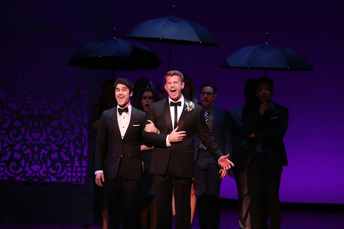 Two men in tuxedos stand arm-in-arm, singing on stage with expressive faces. Behind them, people hold black umbrellas, and the background is lit with purple lighting and ornate patterns.
