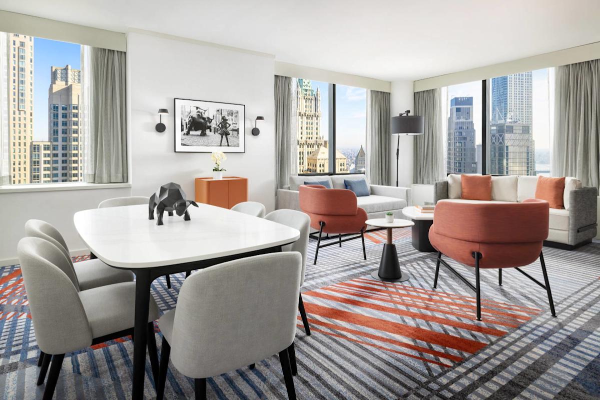 A modern, bright hotel suite features a dining table with chairs, a living area with sofas and orange chairs, large windows with city views, and modern decor including a black elephant figurine and framed artwork.