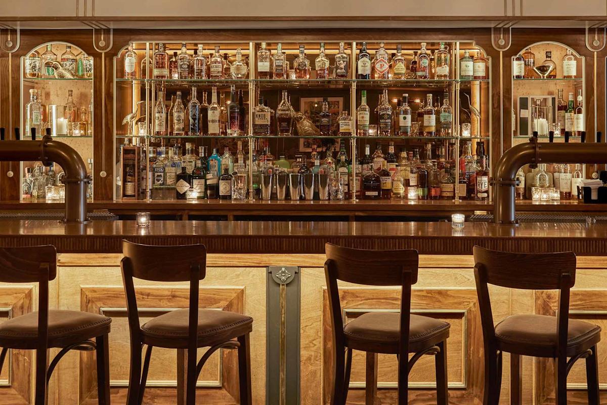 A warmly lit bar with four wooden barstools, a polished counter, and shelves behind displaying various bottles of liquor, cocktail glasses, and small lit candles.