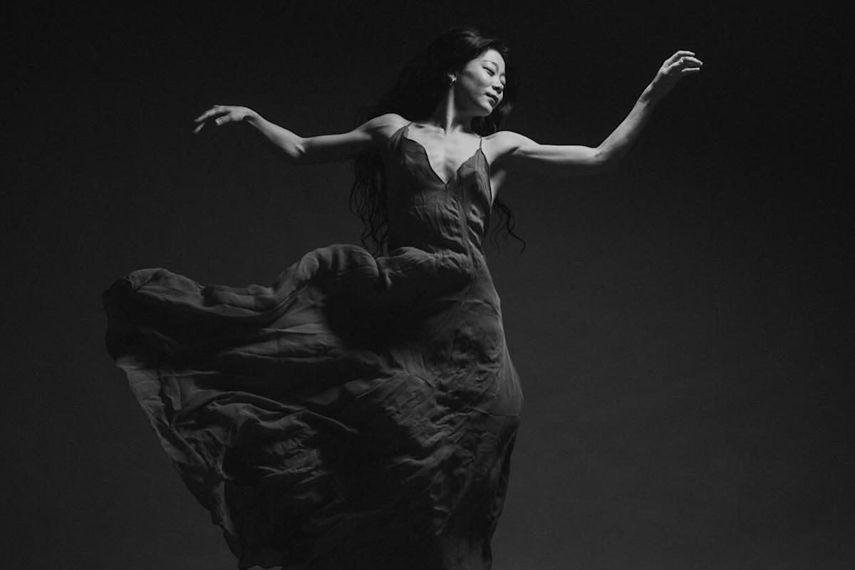A black and white photo of a dancer gracefully moving. She is wearing a flowing dress that swirls around her, with arms elegantly extended. Her expression is focused and serene against a dark background.
