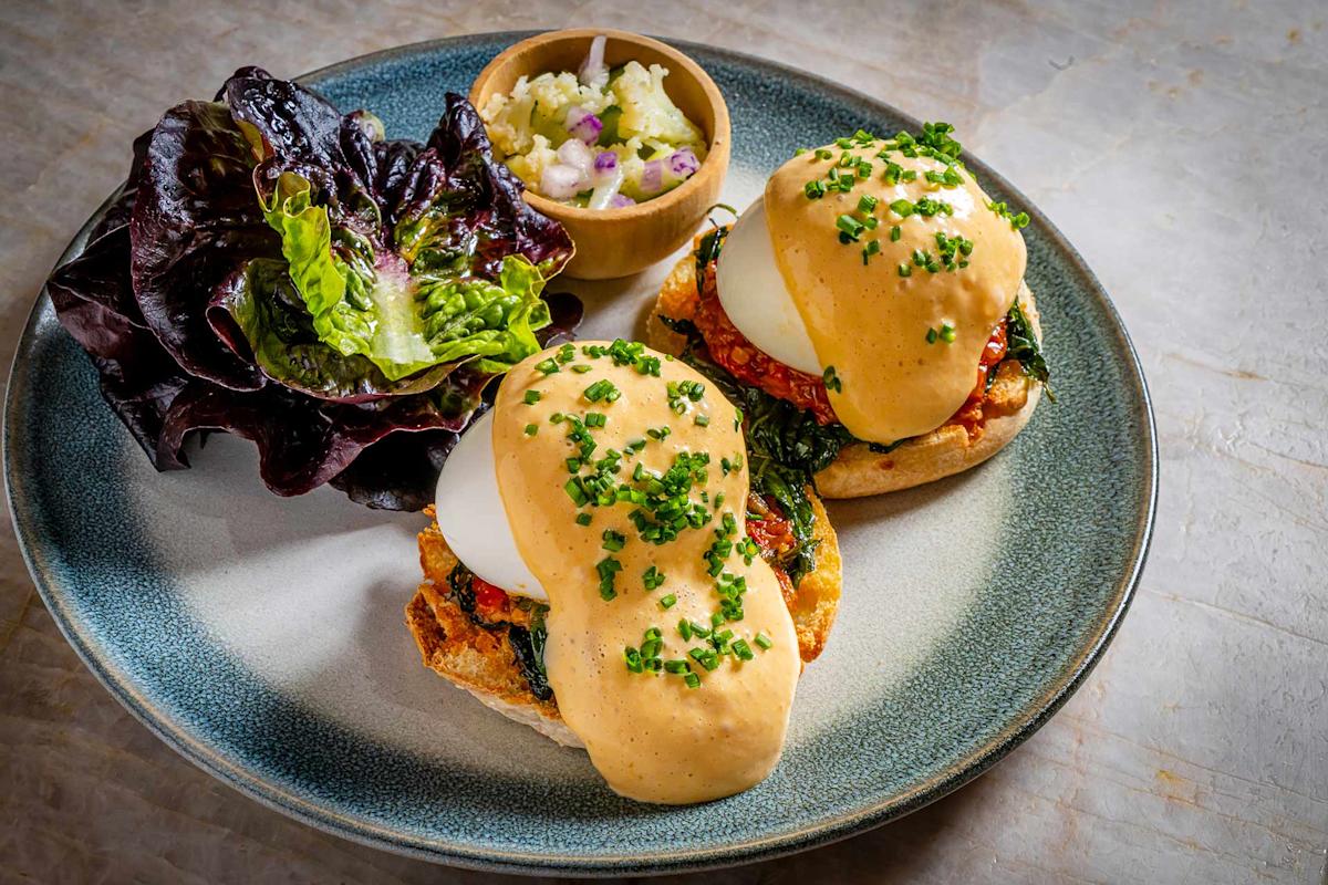 A plate with eggs Benedict topped with hollandaise sauce and chopped chives on English muffins, served with a leafy green salad and a small bowl of potato salad.