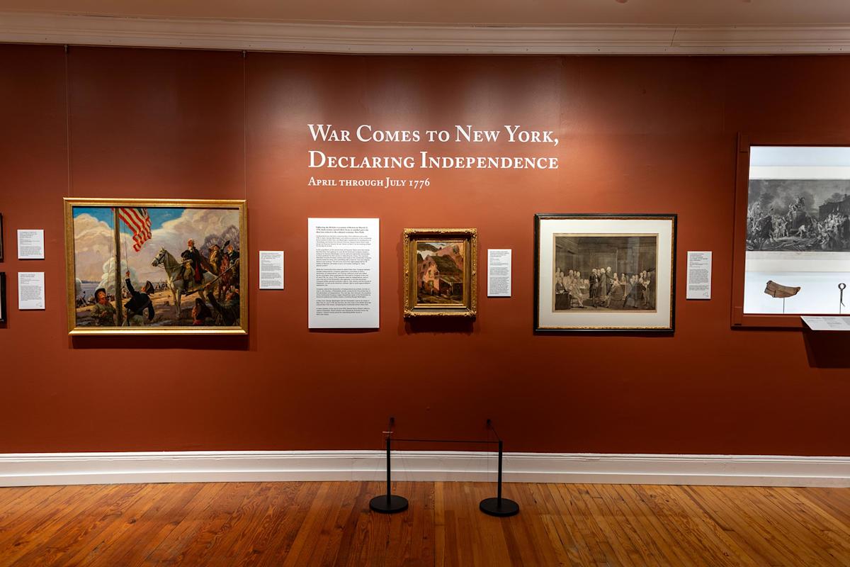 Museum exhibit with brown walls displaying paintings, historical text panels, and a horn. Text on the wall reads, “War Comes to New York, Declaring Independence. April through July 1776.”.