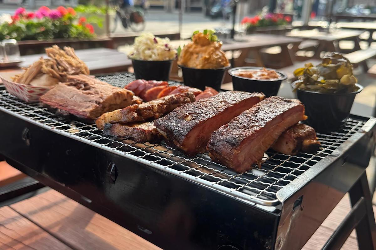 A tray of assorted barbecued meats, including ribs, brisket, and chicken, served with side dishes like coleslaw and pickles, is set on a picnic table at an outdoor restaurant.