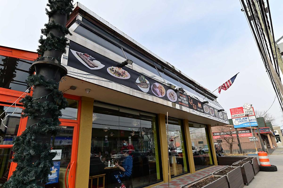 A street view of a restaurant with yellow-framed windows, menu images displayed above, an American flag, and a decorated pole. Signs for other nearby businesses are also visible in the background.