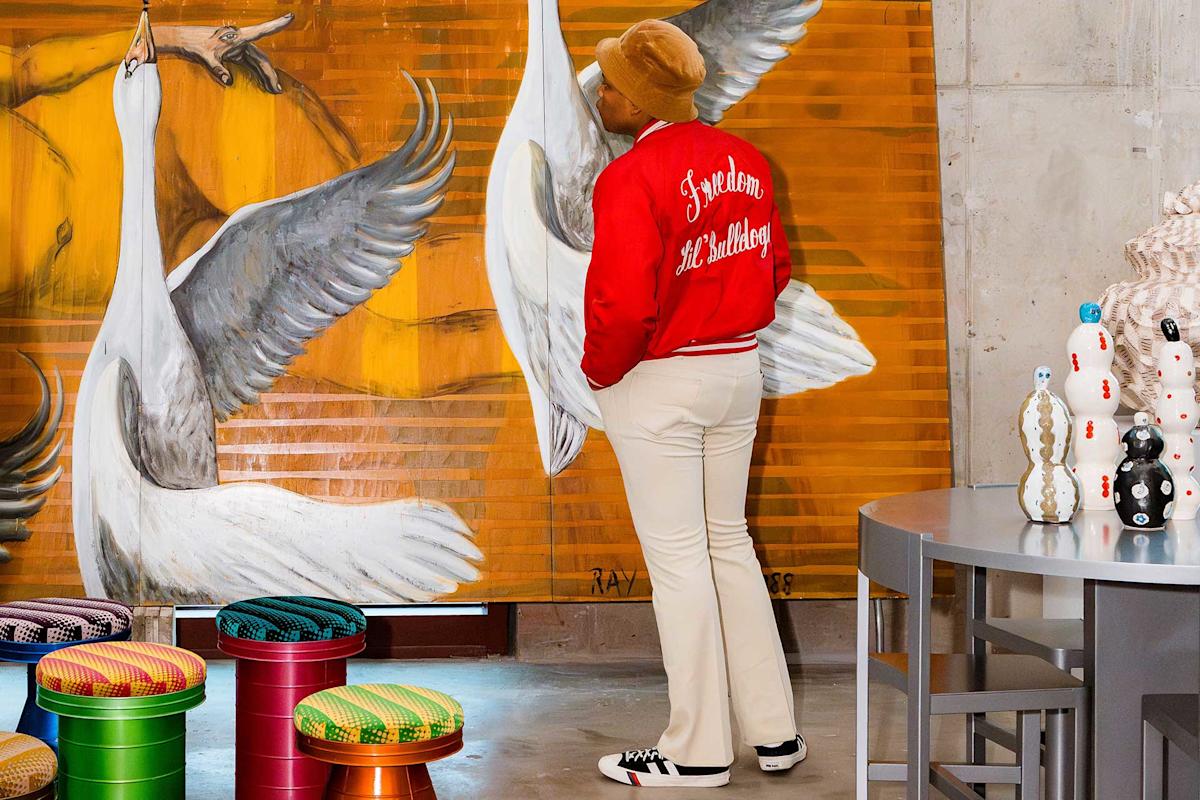 A person in a red jacket, beige pants, and tan hat stands facing a large painting of white cranes. Colorful stools and patterned vases are in the foreground of the modern, artistic space.