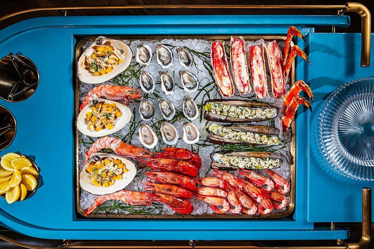 A tray of assorted seafood on ice, including oysters, crab legs, large prawns, clams topped with herbs, and lemon wedges, arranged on a blue cart with plates and utensils on the side.