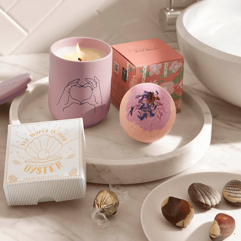 A lit candle in a pink ceramic jar, along with a bath bomb sprinkled with petals and chocolate truffles in the shape of seashells.