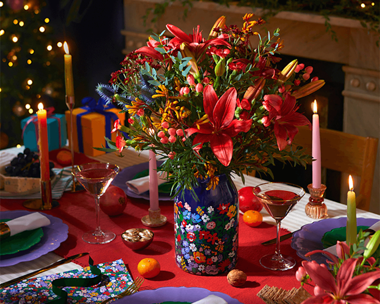A gorgeous arrangement of unusual festive stems – like crimson lilies, blue thistles and golden kangaroo paw – in a patterned vase on a table set for a Christmas dinner party.