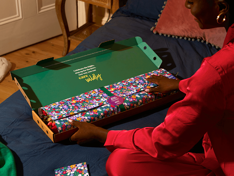 A dark-skinned woman in red silk pyjamas opens a Bloom & Wild letterbox package, to reveal flower stems wrapped in paper and ribbon. The paper is printed with a vibrant Liberty design.